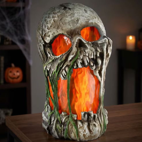 A Halloween skull-shaped lamp with glowing orange fake flames and green vine details, displayed on a wooden table in a dimly lit room.