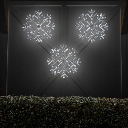 Multiple cool white LED snowflake Christmas decorations glowing outdoors above evergreen shrubs at night, creating a festive winter light display.