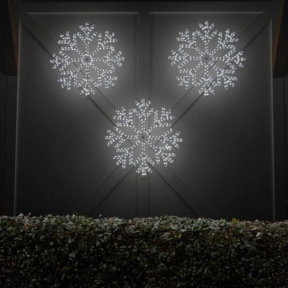 Multiple cool white LED snowflake Christmas decorations glowing outdoors above evergreen shrubs at night, creating a festive winter light display.